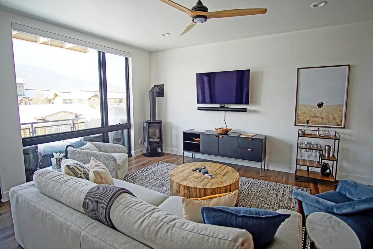 A welcoming living area with large windows, wooden coffee table, plush sofas, and a TV. The space is bathed in natural light and is arranged to foster socializing, relaxation, and entertainment, with outside views easily accessible from the comfort of the indoors.