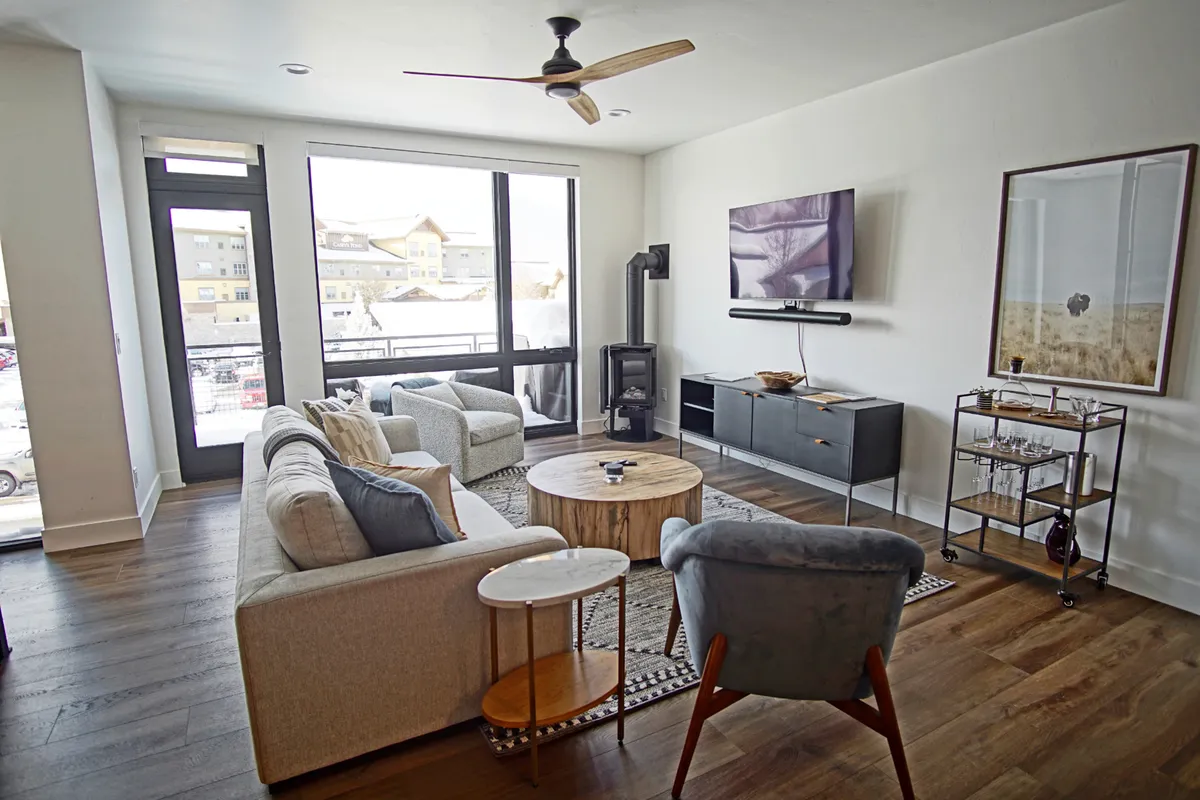 A cozy, modern living area with large windows flooding the space with natural light. A round wooden coffee table sits at the center, surrounded by comfortable seating, artwork, an entertainment center, and views of the outdoors.