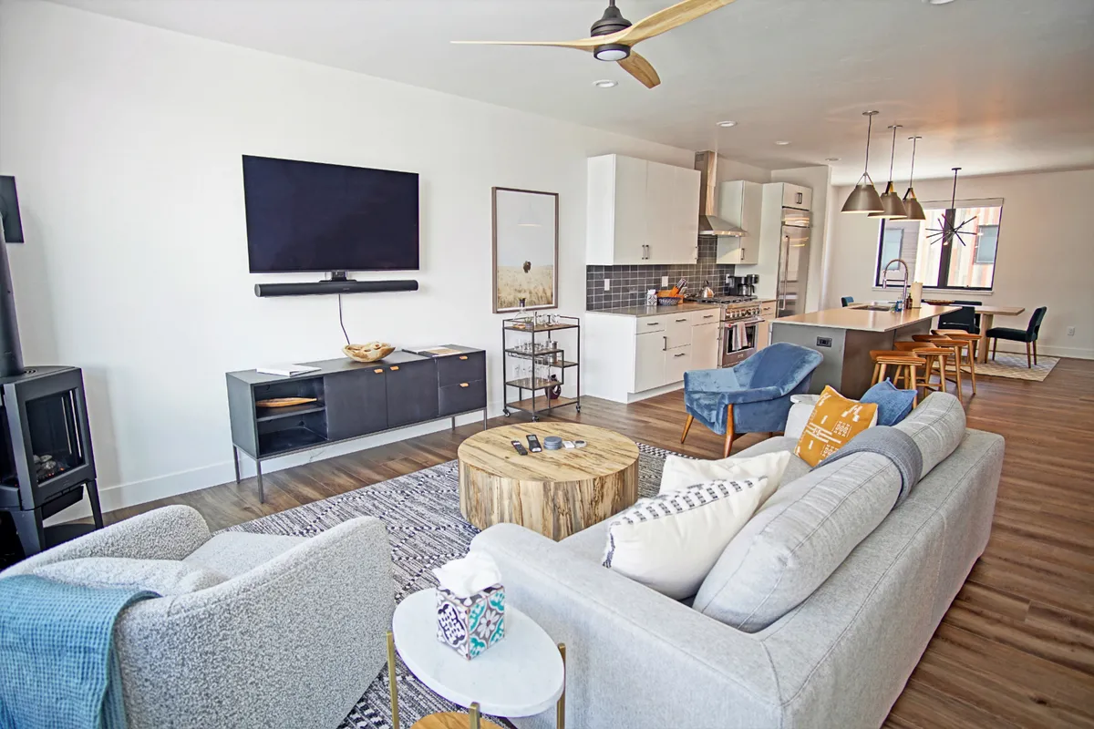 A wide-angle view of an open-plan space that seamlessly combines a large television, wooden coffee table, comfortable seating, kitchen, and dining area. The setting is warm, modern, and flexible—ready for movie nights, shared meals, or morning coffee routines.