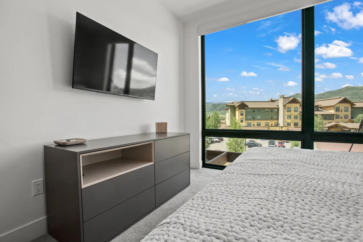 A serene bedroom with a large window that frames a stunning view of the mountains. The room is decorated with textured bedding and a modern TV, hinting at a relaxing retreat after a day spent outdoors.