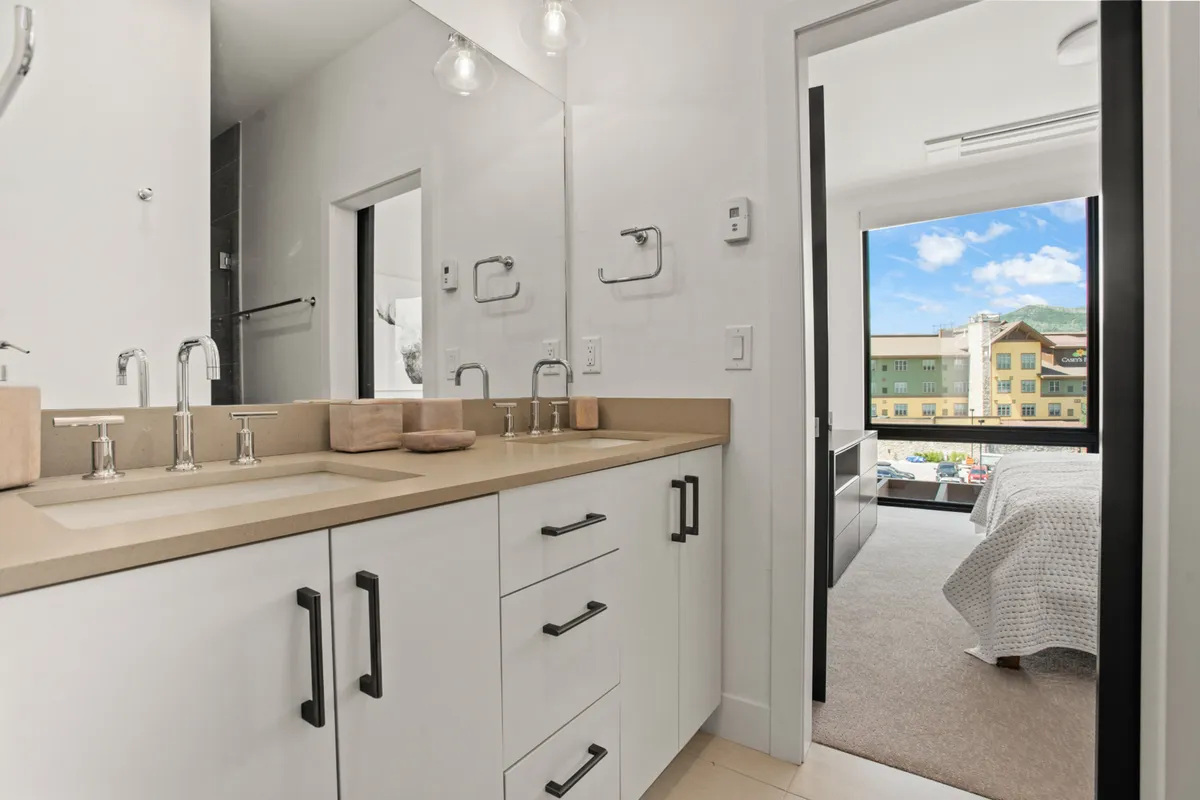 A spacious bathroom with a double sink vanity, large mirror, and window offering a view into the adjacent bedroom. The room is brightly lit with natural light, and the thoughtful design enhances the daily routine with both functionality and comfort.