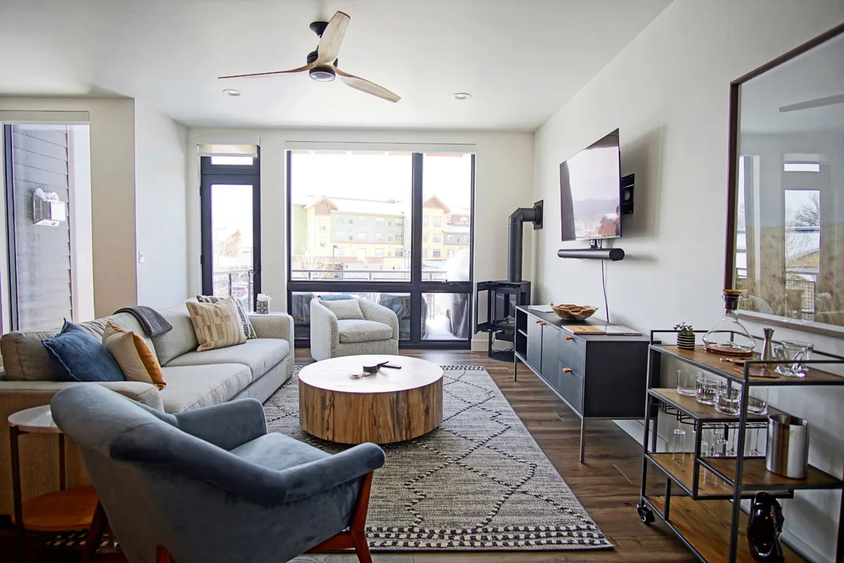This image features a spacious living area with large windows that provide panoramic views of the surrounding landscape, a wooden coffee table, and comfortable seating. The abundance of natural light and clear sight lines to the outdoors evoke a strong sense of connection to nature and the local scenery.
