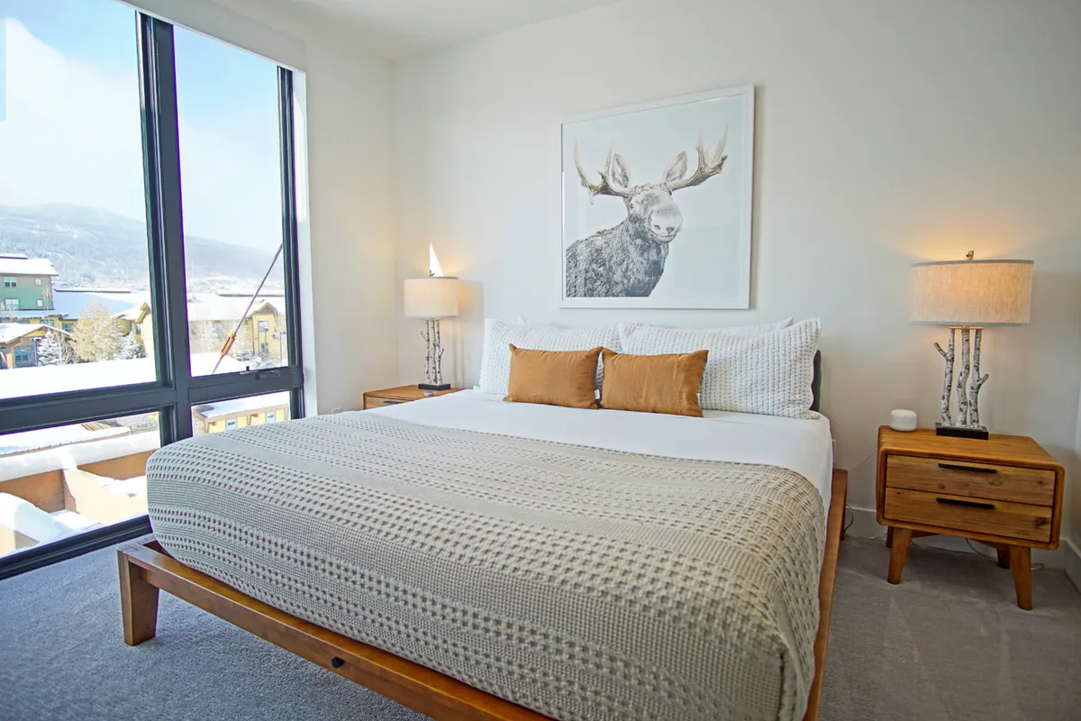 A serene king bedroom suite with a large, inviting bed adorned with crisp white linens, moose artwork above the headboard, and stylish bedside lamps. Natural light filters softly into the room, creating a tranquil retreat ideal for restful sleep or private relaxation.