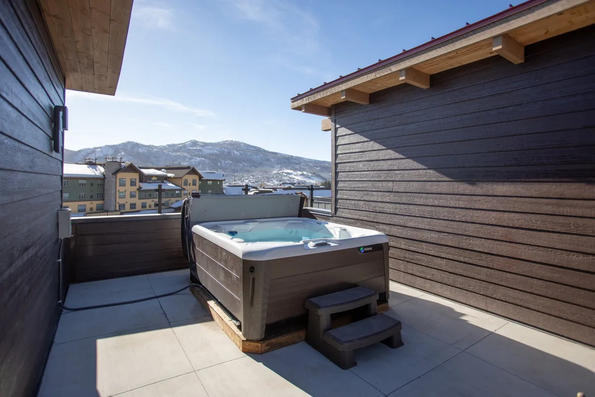 A tranquil rooftop scene focused on a hot tub overlooking expansive mountain ranges, with surrounding buildings visible in the distance. The setting is peaceful and luxurious, perfect for unwinding after a day of adventure or taking in the sunset with friends or family.